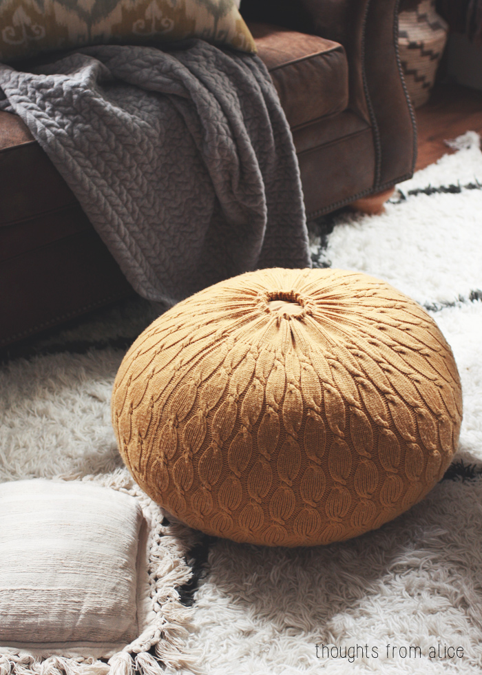 Round tan knitted pouf on a shaggy rug beside a brown sofa with a gray knitted throw.
