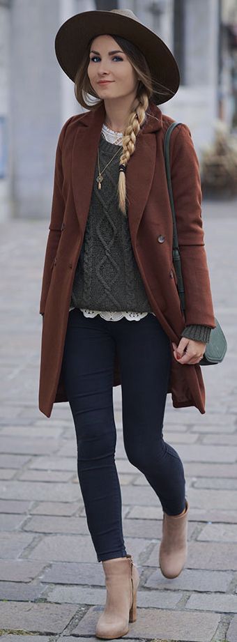 Stylish woman walking city street in a rust-brown coat, gray knit sweater, dark jeans, tan boots, and a wide-brim hat.
