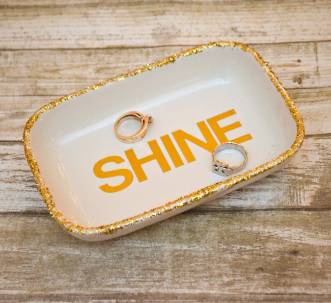 White ceramic tray with a gold glitter rim displaying SHINE in orange, with two rings resting on it on a wood surface.
