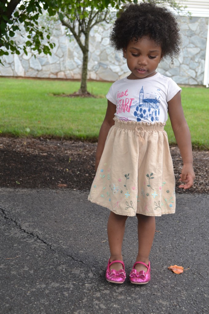 Young girl standing on a paved path in a beige embroidered skirt, pink sparkly shoes, and a white graphic tee.