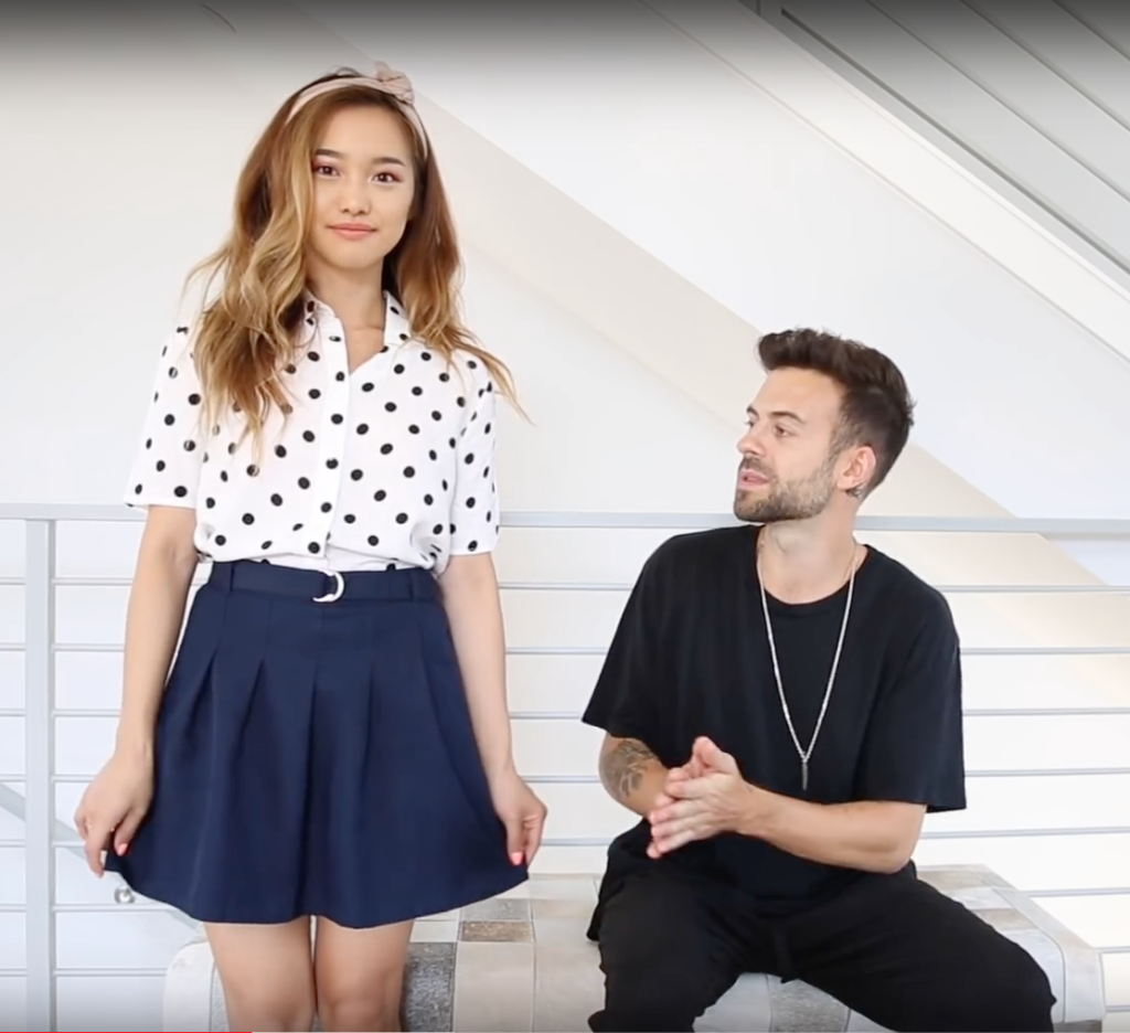 Young woman in a white polka-dot blouse and navy pleated skirt stands beside a seated man in a black T-shirt.