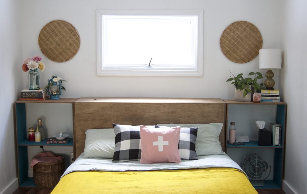 Cozy bedroom with a wooden bed, plaid pillows, yellow quilt, built-in blue shelves, potted plant, and round wicker wall accents.
