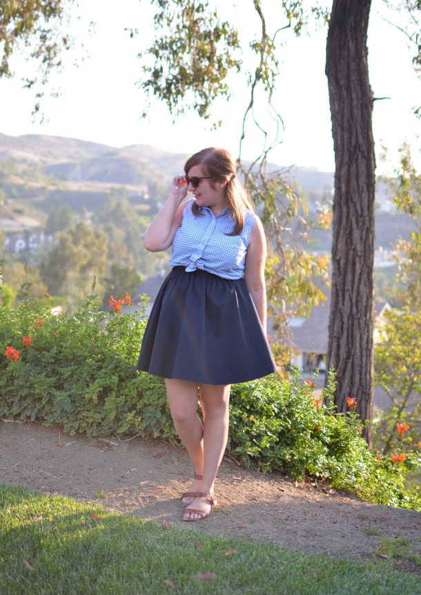 Woman in a blue checkered sleeveless blouse and navy A-line skirt posing on a dirt path in a park with trees and hills.