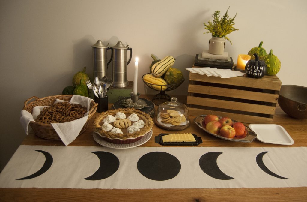 Autumn table spread featuring pies, cheese, crackers, pretzels, apples, and gourds along a moon-phase runner.