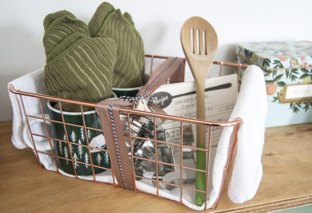 Copper wire basket on a wooden surface filled with green knitted towels, a wooden spatula, a wrapped gift, and assorted kitchen tools.