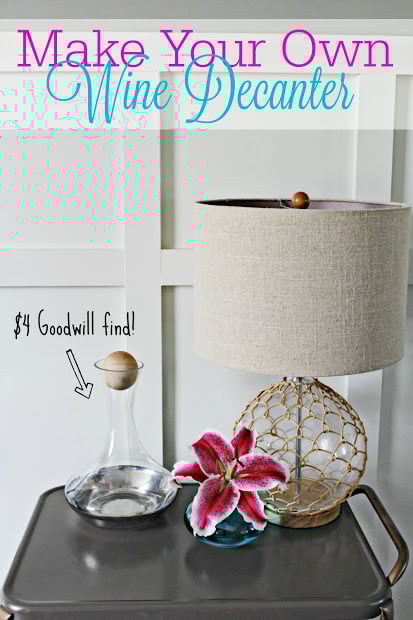 Thrifted glass wine decanter on a small table beside a beige lampshade and pink flower.