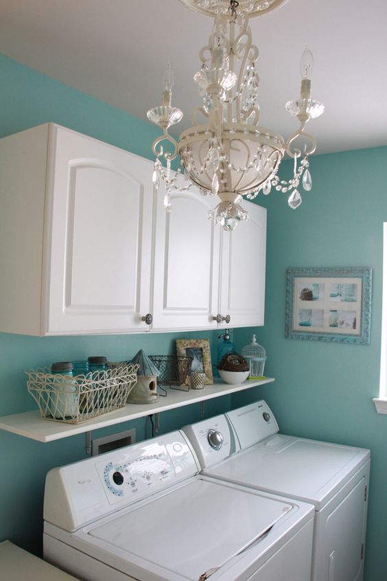 Bright laundry room with white cabinets, teal walls, a crystal chandelier, and a front-loading washer and dryer.