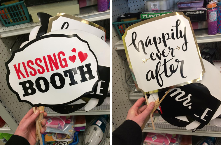Two handheld signs on sticks in a store aisle: 'KISSING BOOTH' on the left and 'happily ever after' on the right.