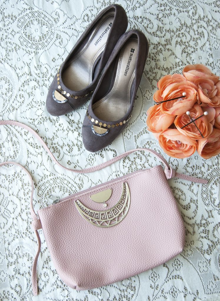 Pink textured crossbody bag with gold-tone half-moon clasp, gray flats, and a peach bouquet on lace fabric.