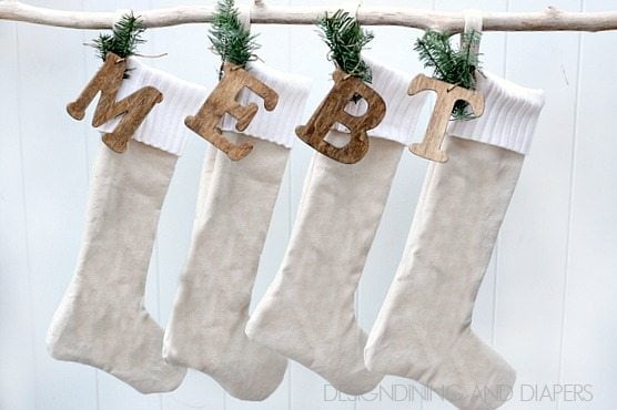 Four beige stockings with white cuffs hang from a rope, with pine sprigs and wooden cutout letters above them.