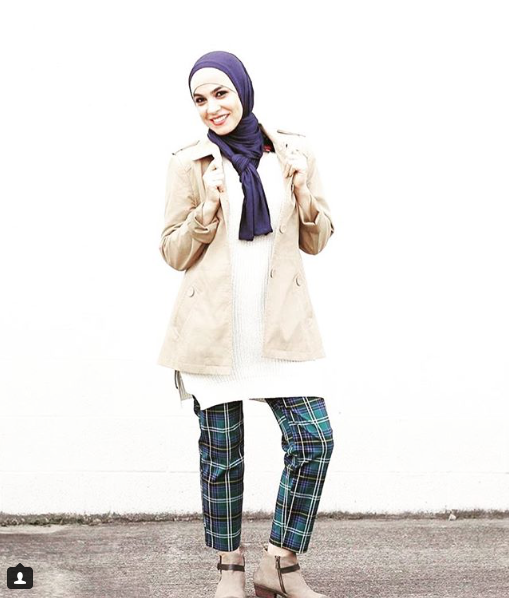 Smiling woman in a purple hijab, beige coat, white sweater, and plaid blue-green pants.