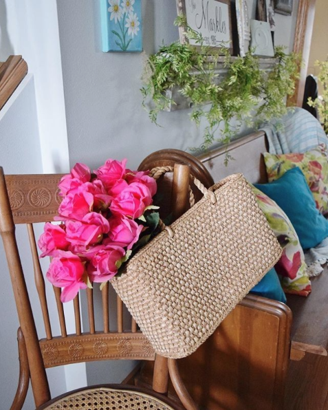 Woven straw handbag with bright pink roses resting on a wooden chair in a cozy living room.