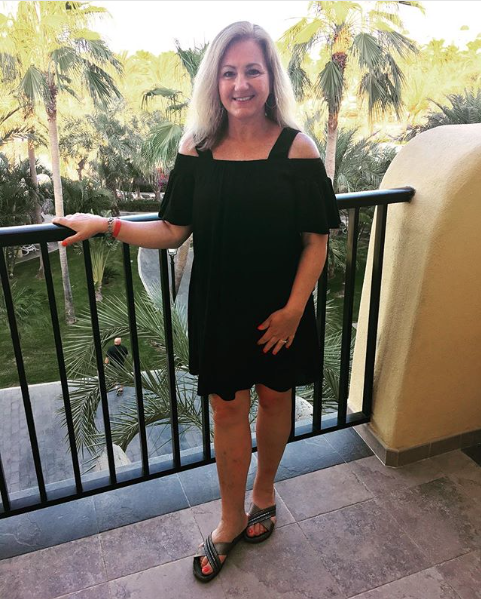 Woman in a black off-shoulder dress standing on a balcony with palm trees behind her.
