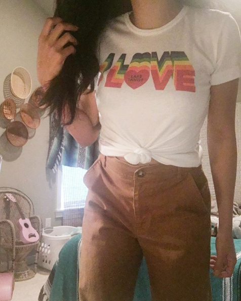 Person wearing a knotted white T-shirt with a rainbow LOVE graphic and brown pants, standing in a cozy room with guitars.