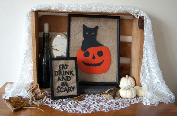 Halloween-themed display in a wooden crate: framed pumpkin-cat art, a spooky sign, small white pumpkins, and lace fabric.
