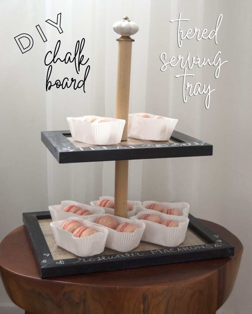 Two-tier wooden serving tray with pink macarons in white liners and chalkboard-edged rims.
