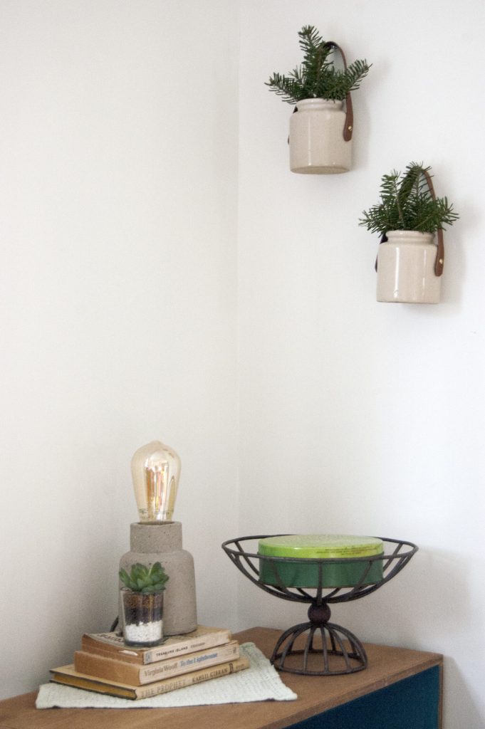 Minimalist corner with two beige hanging planters, greenery, a concrete lamp, a succulent on books, and a wire bowl.
