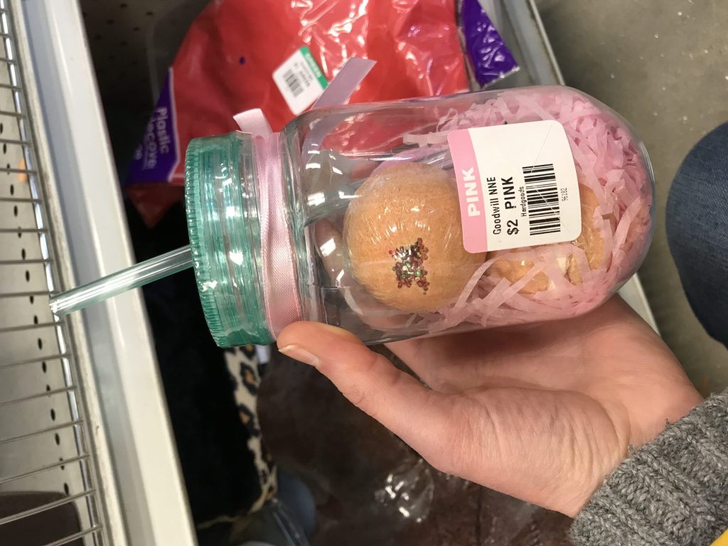 Glass jar with pink filler and a peach bath bomb, held by a hand; Goodwill Pink tag on the label.