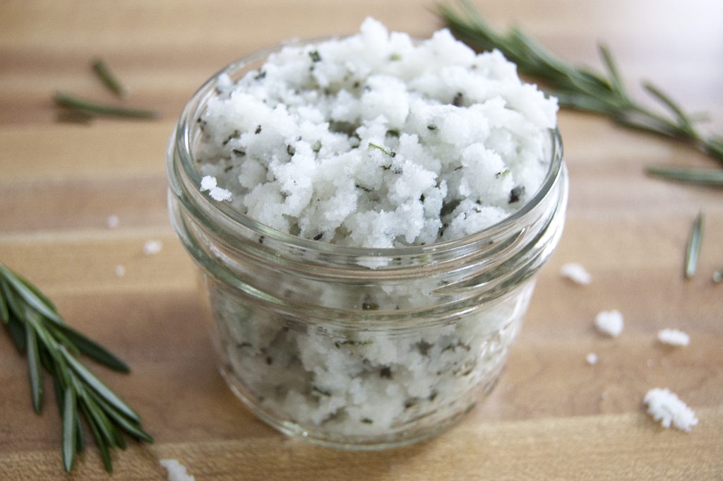 Coarse rosemary-infused sea salt in a glass jar on a wooden surface.