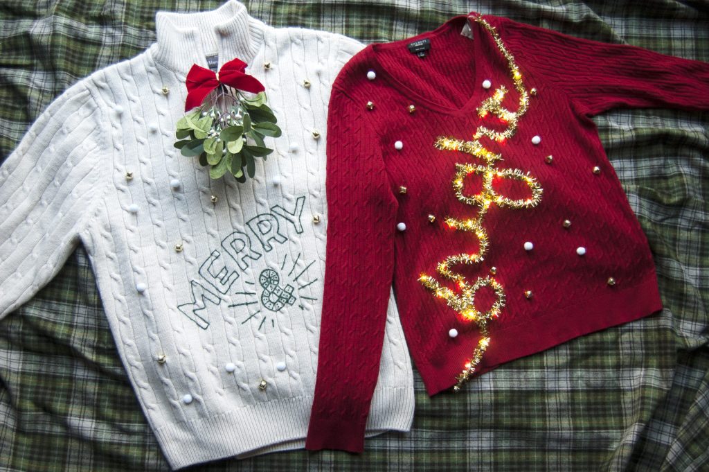 Two Christmas sweaters on a plaid blanket: white cable knit with 'MERRY' and greenery bow, red knit with gold tinsel spelling 'Noel'.