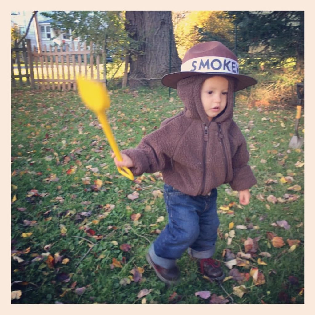 Young child in a brown hooded jacket and hat with a 'SMOKE' band, holding a yellow toy shovel in a leaf-strewn yard.