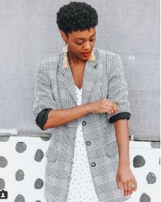 Woman in a gray plaid blazer over a white polka-dot dress, rolling up her sleeve against a gray wall.