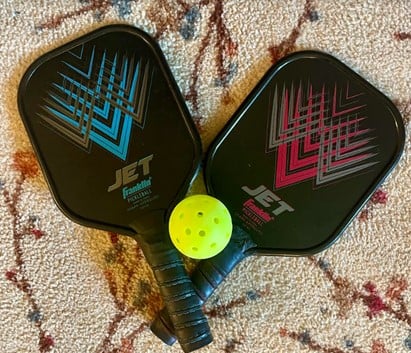 Two black pickleball paddles with turquoise and pink chevron patterns and a neon yellow perforated ball on a patterned rug.