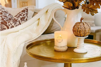 Cozy living room scene with a candle on a gold tray, pumpkin decor, and a knit blanket beside floral cushions.