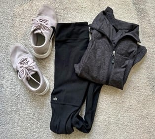 Light beige sneakers, black leggings, and a gray zip-up hoodie laid out on a beige carpet.
