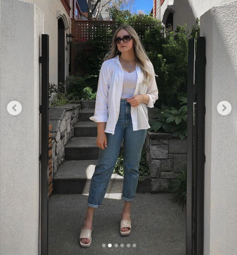 Woman in white shirt and jacket, jeans and sandals, standing outdoors between stone pillars with greenery.