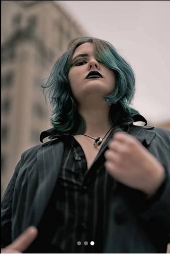 Women with a short blue hair and black lipstick wearing a black jacket and black and grey stripped blouse. 