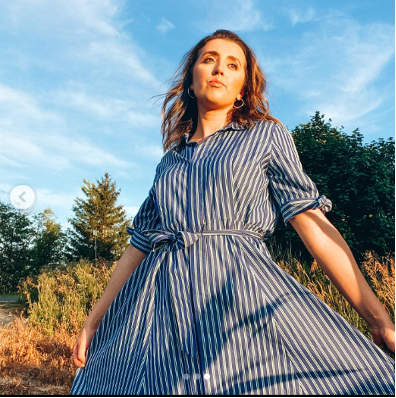 Woman in striped blue dress standing in a sunlit field with trees under a clear blue sky