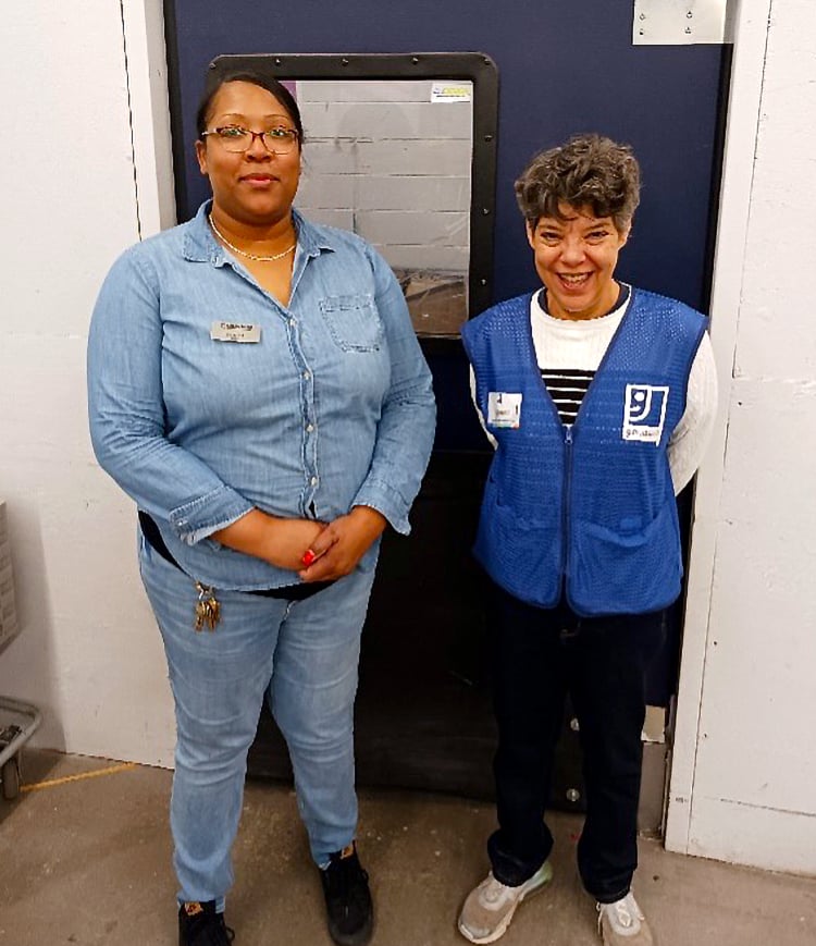 Two women stand indoors by a large service door; one wearing a denim shirt and jeans, the other in a blue safety vest.