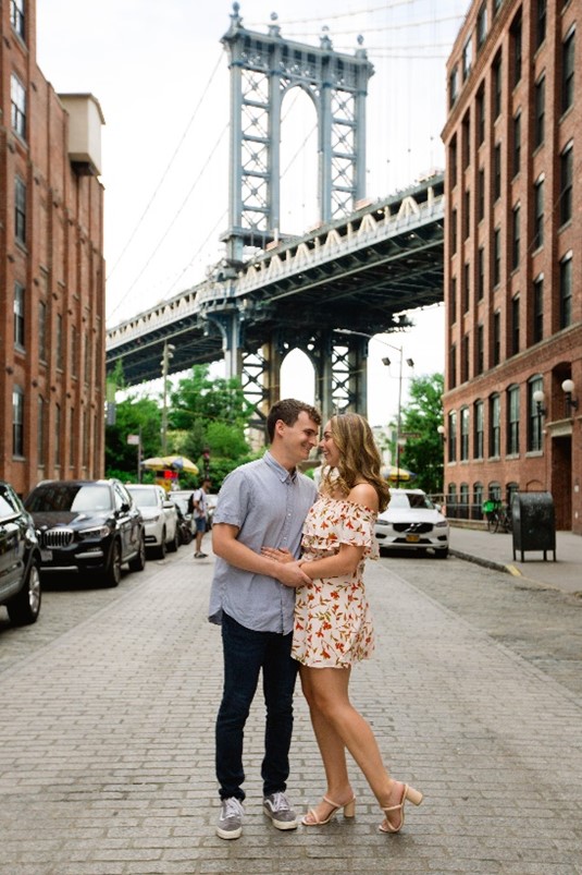 Couple embracing in a cobblestone street under a large suspension bridge, flanked by brick buildings.