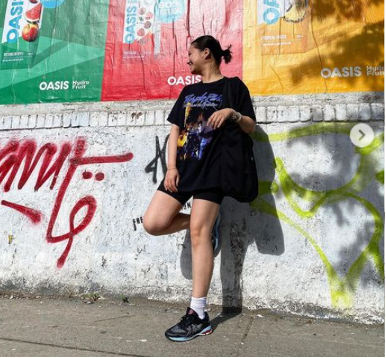 Young person leaning against a graffiti-covered wall with colorful Oasis banners