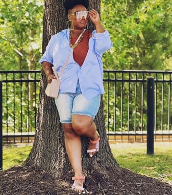 Woman leaning against a tree in a park, wearing oversized blue shirt, denim shorts, sandals, sunglasses, and a beige purse.