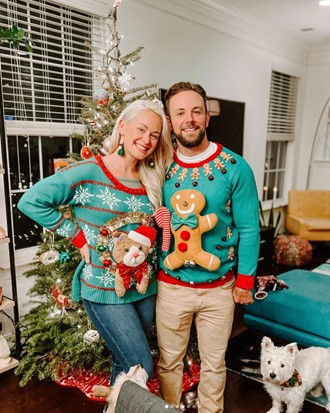 Smiling couple in festive teal Christmas sweaters posing beside a decorated tree with a small white dog nearby.