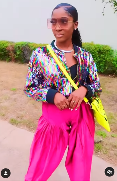 Person in a multicolor sequin jacket, hot-pink pants, black top, sunglasses, and neon yellow crossbody bag, posing outdoors.