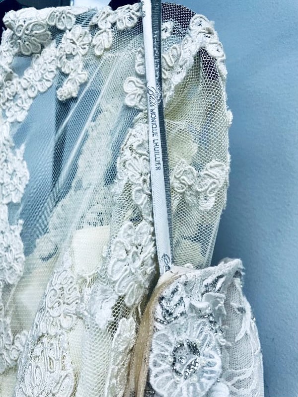 Delicate white lace wedding dress with floral embroidery on a hanger against a blue wall.