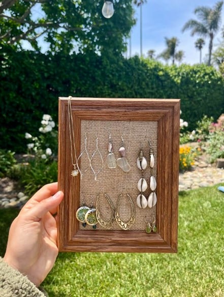 Hand holds wooden frame with burlap jewelry display of assorted earrings in a sunny garden.