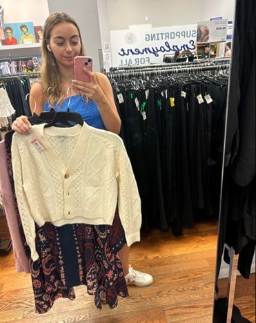 Young woman in a clothing store holds a cream knitted cardigan while taking a mirror selfie.