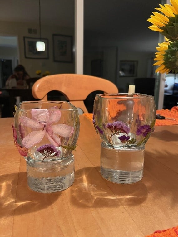 Two clear glass cups filled with water and decorative flowers sit on a wooden table, with sunflowers in the background.