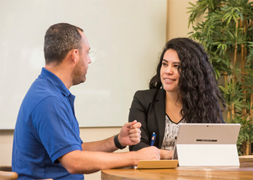 Man in a blue polo and a woman with curly hair discuss across a desk with a tablet in a bright office.