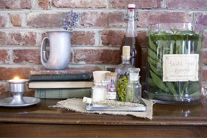 Rustic still life on a wooden table before a brick wall: candle, pitcher, books, cork bottle, and jars of spices.