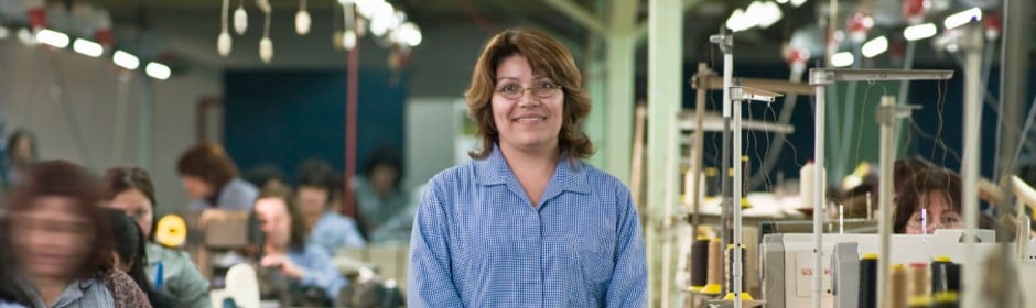 Smiling woman in a blue checkered shirt stands in a busy sewing workshop with machines and workers in the background.