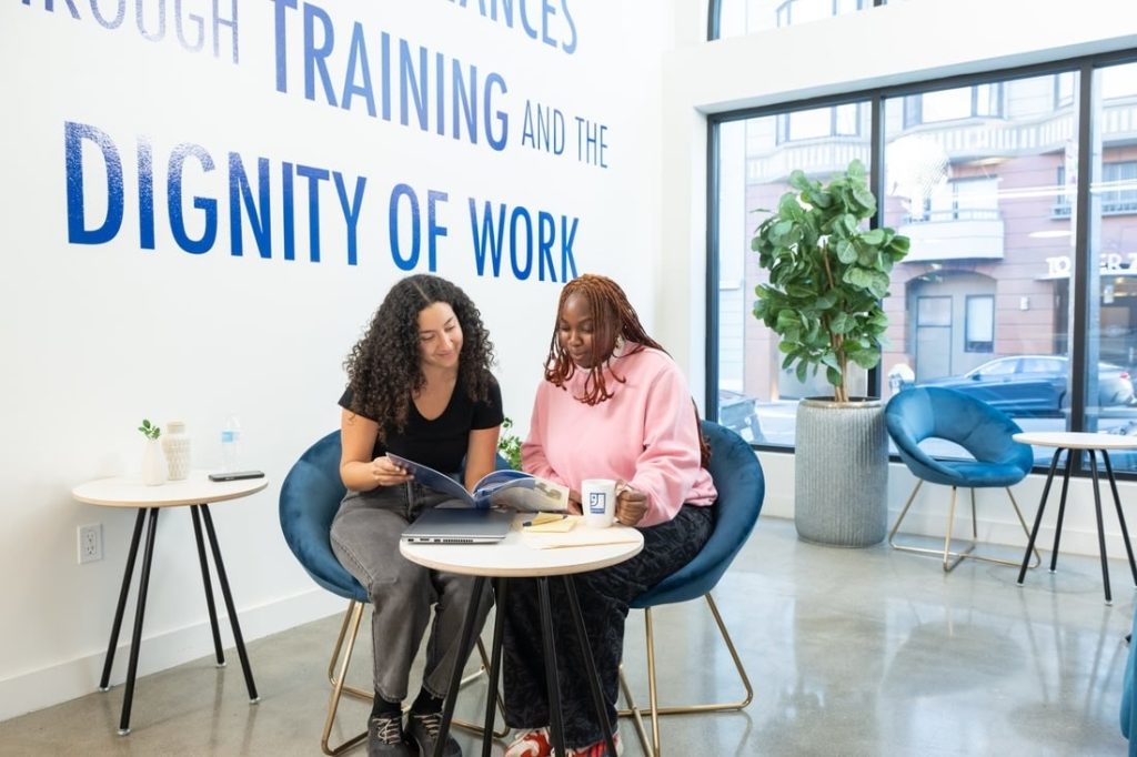 Two women collaborate at a modern coworking space with bold wall text reading DIGNITY OF WORK.