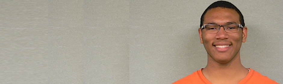 Smiling young man in an orange shirt and glasses against a light gray wall.