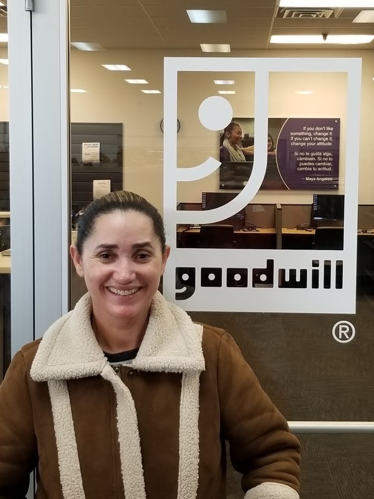 Smiling woman in a brown shearling coat stands in front of a glass door bearing the Goodwill logo.
