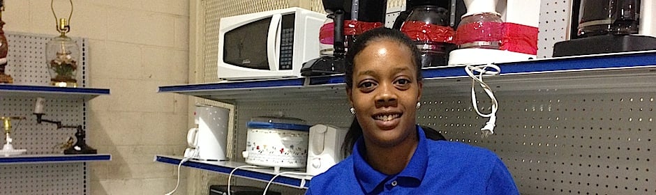 Smiling store associate in a blue polo in a kitchen appliance aisle with microwaves and blenders on shelves.