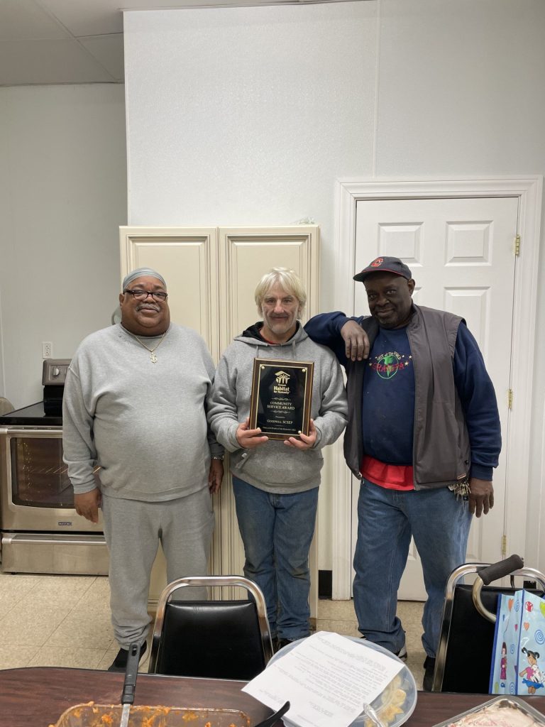 Three men stand indoors in a kitchen-like room; the center man holds a plaque award while the others smile.